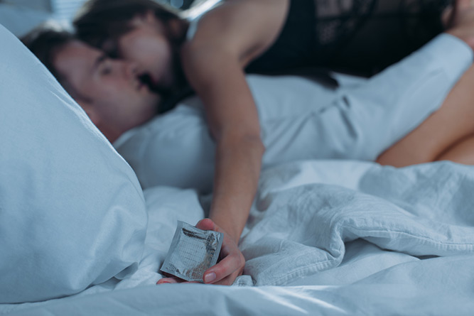 woman on top of a man in bed, holding a condom in her hand